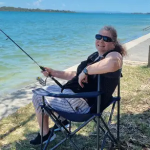 The LevelUp NDIS Support Worker is enjoying fishing while sitting in a folding chair with a fishing rod by the water's edge.