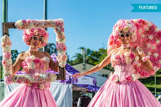 Two women wearing pink dresses and floral headpieces smiling at a Queensland Garden Expo.