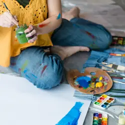 A woman in a yellow top and denim pants paints with colorful paint and brushes on a canvas for LevelUp Creative Art Therapy.